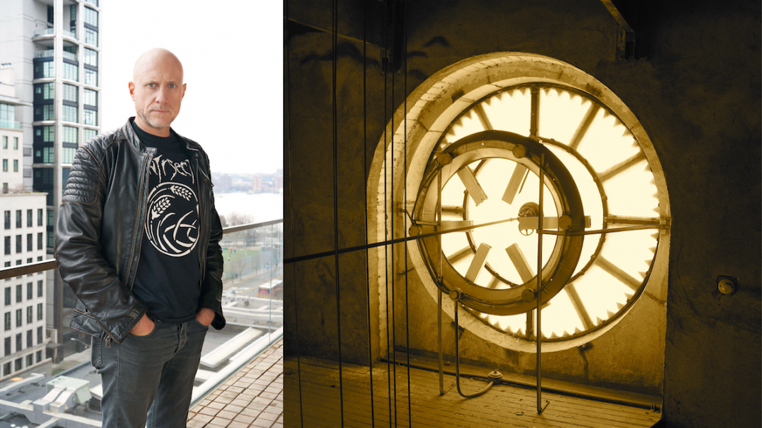 Bald white male wearing black graphic t-shirt and leather jacket on a balcony overlooking an urban landscape; interior of a large clock in a tower, with gears and rods.
