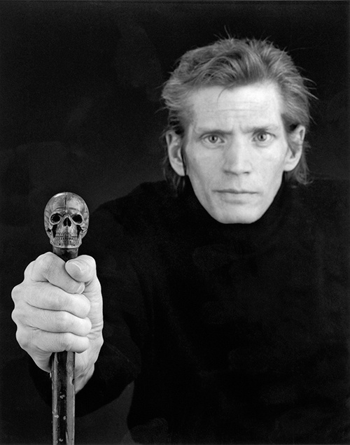 A black and white photograph of a blonde man looking directly into the camera. He is holding a cane with skull on it. He leans forward against a black backdrop, wearing a black coat that fades into background.
