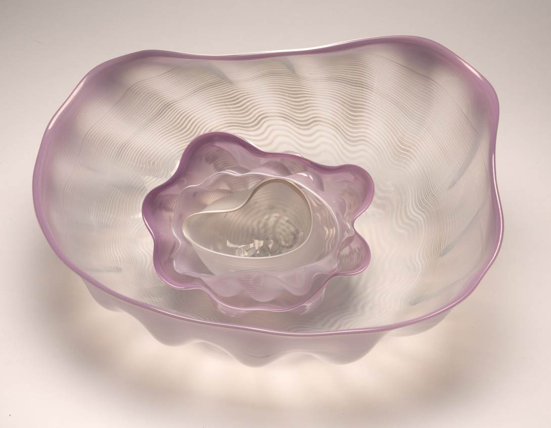 A photograph of 4 wavy clear glass bowls that have edges tipped with pink. They are nested within each other and resemble a flower.