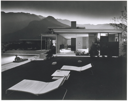 A black and white photograph of a modern home's backyard with two chaise loungers and a table. There is a lit pool with a person on a mat lying beside the pool. In the near distance are hills with light reflecting off the mist of the evening air.