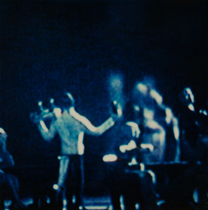A blurred photograph of figures, dimly lit by a blue glow. The most visible figure appears to be a waiter—possibly carrying a serving tray with two drinks in one hand, a third drink in the other, and a towel or napkin hangs from its back pocket.
