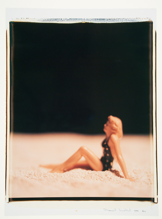 A polaroid photograph of a blonde female doll or sculpture in a black, polkadot bathing suit sitting on sand. The black background is slightly blurred, and her arm and the sand are more focused.