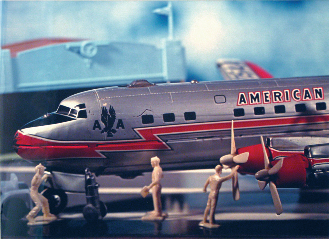 A photograph of a replica of the front half of an America Airlines plane with replicas of 3 airport maintenance workers. The background is out of focus showing an airplane hanger with a flag.