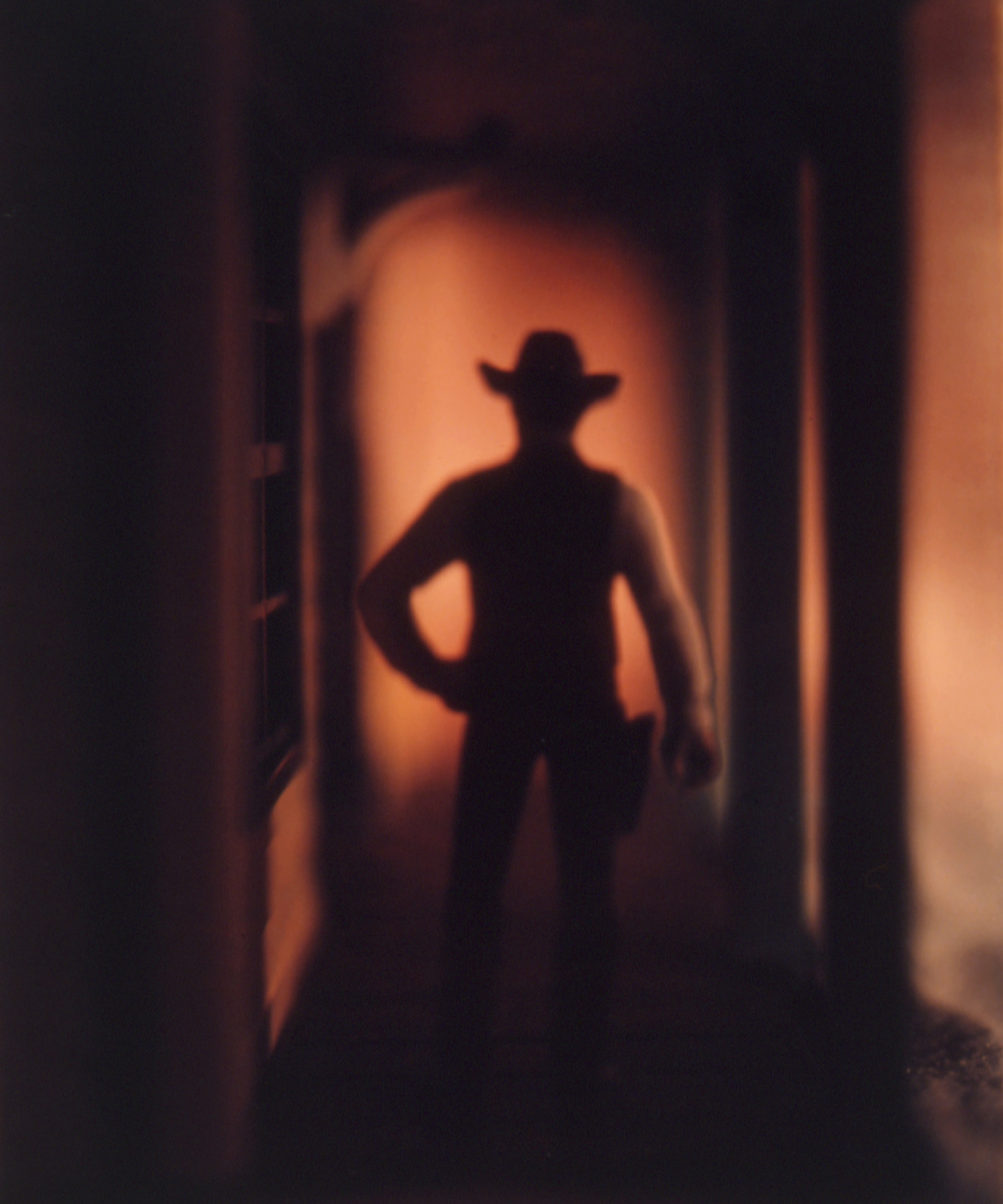 A German-designed cowboy figure—saved from a set given to the artist by his parents—stands in a hallway to scale. Its hand on one hip, a holster on the other. The scene is blurred, and a warm orange glow casts the figure and pillars in shadows.