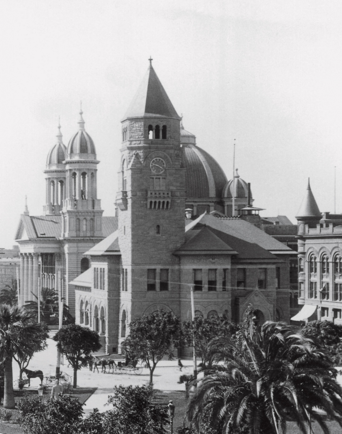 A historical black and white photograph of a brick building that was originally a post office before it became the San José Musuem of Art. There are horse-drawn carriages in the foreground. Palm trees overtake the view.