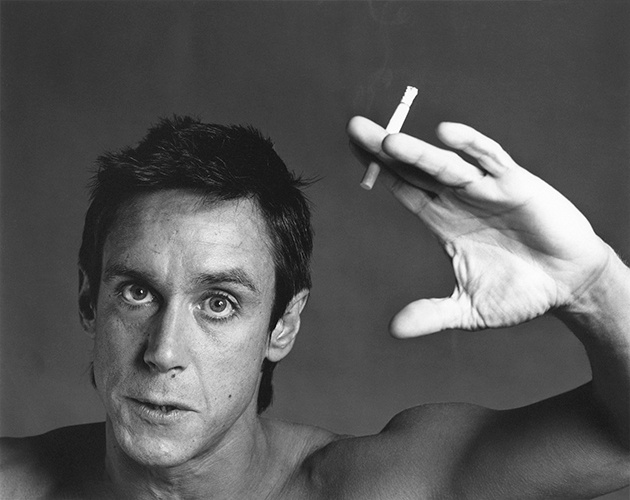 A black and white photograph of the punk singer Iggy Pop. He is shirtless, holding a halfway smoked cigarette with a lot of ash. His hand is slightly open and directed towards the camera. He is postured, leaning in, his mouth slightly open.