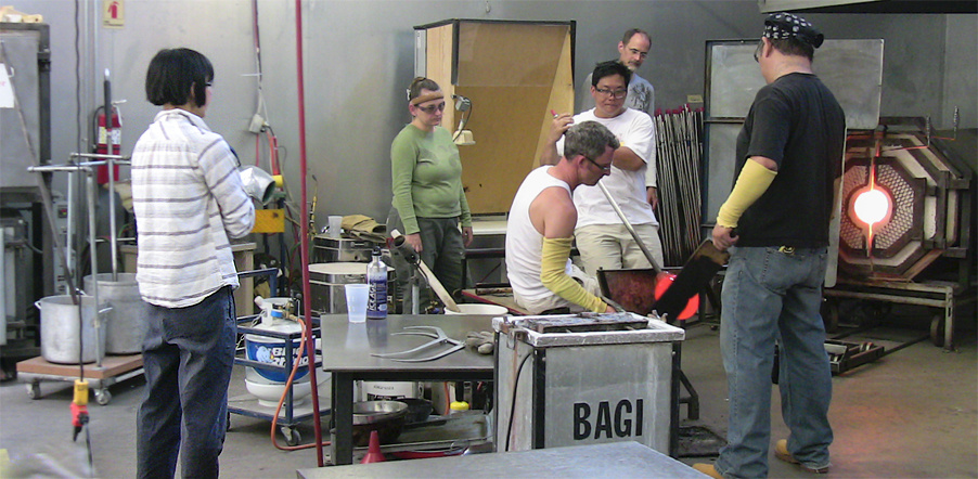 Six individuals working in a glass studio. Two of the artists are engaged in a process while the other four observe at a slight distance. 