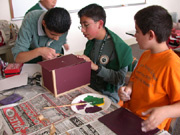 Three boys stand around a large table that is covered in newspaper, fully engaged in building and painting a birdhouse. There is a paper plate with paint colored green, yellow, purple and black. 