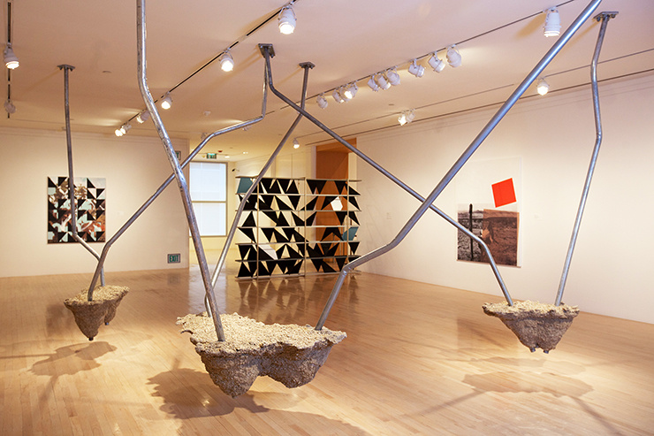 A large sculpture made of bent metal tubes connects suspended masses of hardened cement. The tubes are mounted into the gallery ceiling with hardware. The lit sculpture creates shadows. In the background, a sculpture of black triangles acts like a screen within the space.