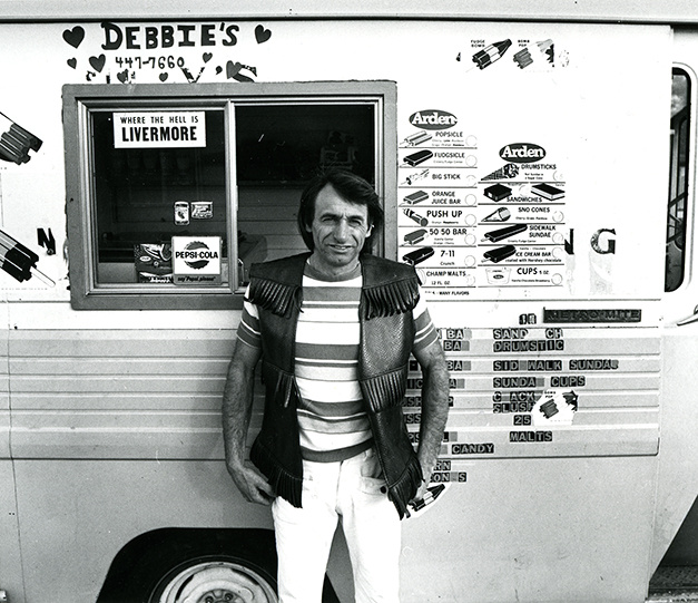 A black and white photograph of a man wearing a striped shirt and fringed leather vest standing in front of a Debbies ice cream truck. It is covered in stickers that note the price of ice cream treats and the window is open, also partially covered in stickers.