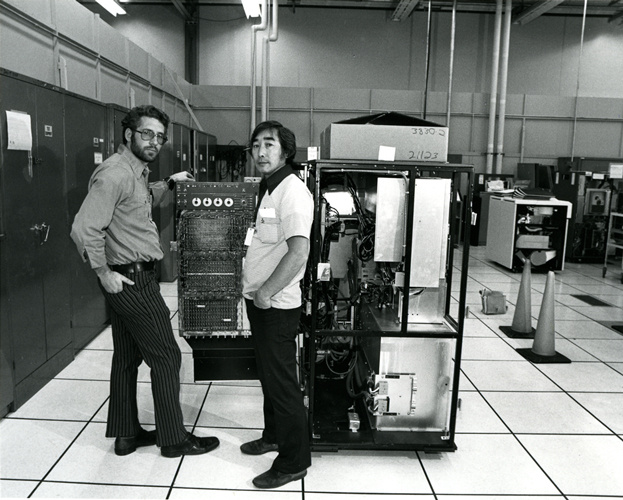 A black and white photograph of two men standing next to a very large computer. They appear to be in a large office space, with traffic cones to the right of them. Next to them is large metal rack is filled with boxes and computer equipment.