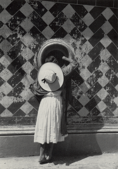 A black and white photograph of a lady in a dress wearing a sombrero on her back is looking into a circle-shaped hole in a wall. The wall is a checkered pattern with some light damages as if something has scraped it.