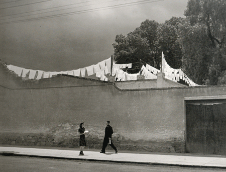 A black and white photograph of two figures in mid-stride. They approach one another on an otherwise empty road. The young woman carries a parcel as though making an offering. The man hurries along, propelled forward by his lengthy gait.