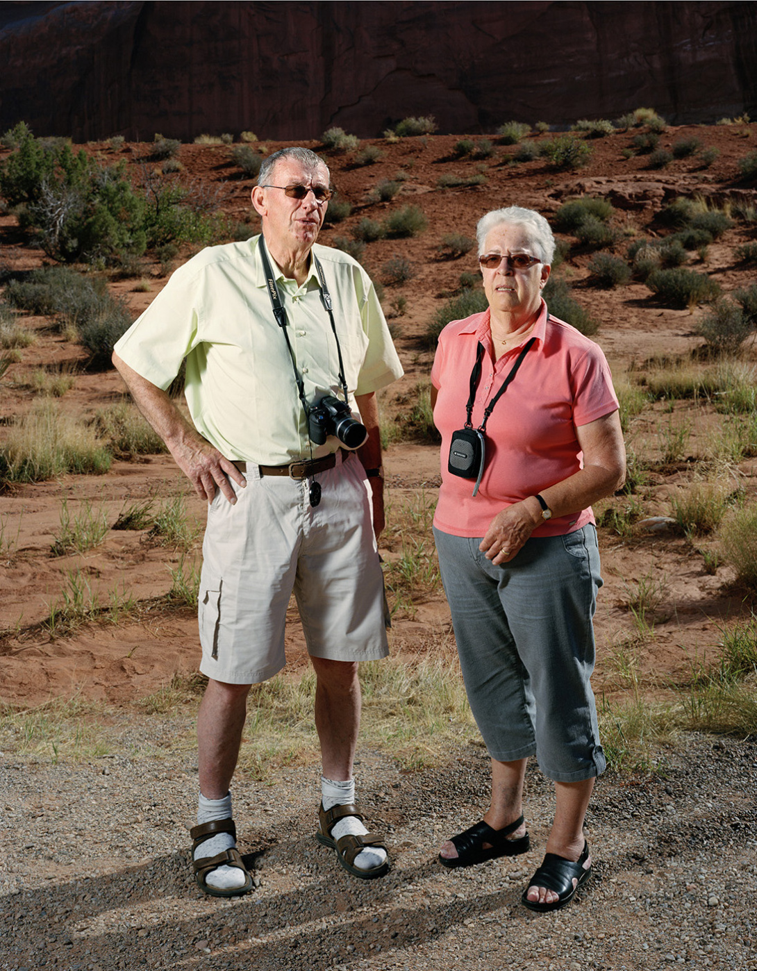 An older man and woman who are dressed for the heat—wearing sunglasses, short sleeved shirts, and sandals.—pose for a photograph at dusk. Each have a camera around their necks. They are in front of canyon with red dirt, rocks, and green vegetation.