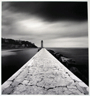 A light house at the end of a very long narrow cement walkway. It is surrounded by a fog, located near a rocky coast. It is either early morning or early evening. Looks cold, and very peaceful place.