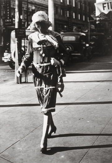 A black and white photograph of a person wearing a long sleeved, long prairie style dress with heels. They are wearing a large sun hat and a mask. Their gender is unknown. They look foreboding walking down a city street with old cars from a past time.