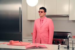 A young man stands behind a kitchen counter. He wears a Pepto Bismol pink suit, shirt, tie, and sunglasses. A cutting board, kitchen utensils, and open book lay on the counter in front of him, all covered in the identical shade of pink. 