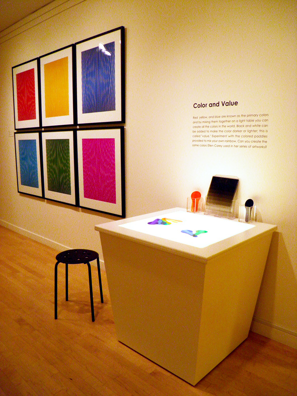 A photograph of a gallery wall. Six pictures of different colors hang in picture frames on the wall next to a table and chairs with colorful objects on it. On the wall is a description of the art piece in black vinyl letters, that reads "color and value."