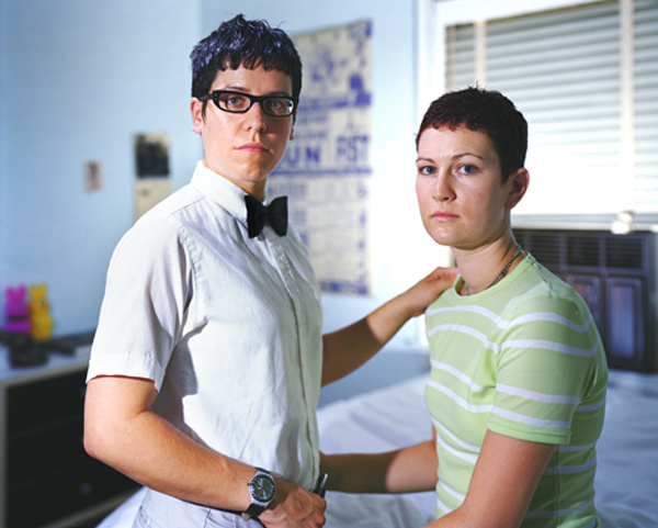 A color portrait of a lesbian couple. They face the camera, faces serious. In the background is a bedroom. One stands while the other sits. They have their arms loosely around each other.