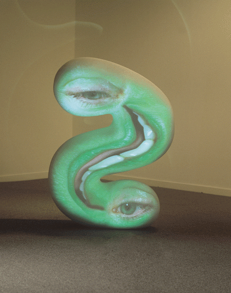 An abstract greenish sculpture that is shaped like a backward "S." It has a vertical smile in the middle—showing teeth and tongue with eyes on each end of the "S."
