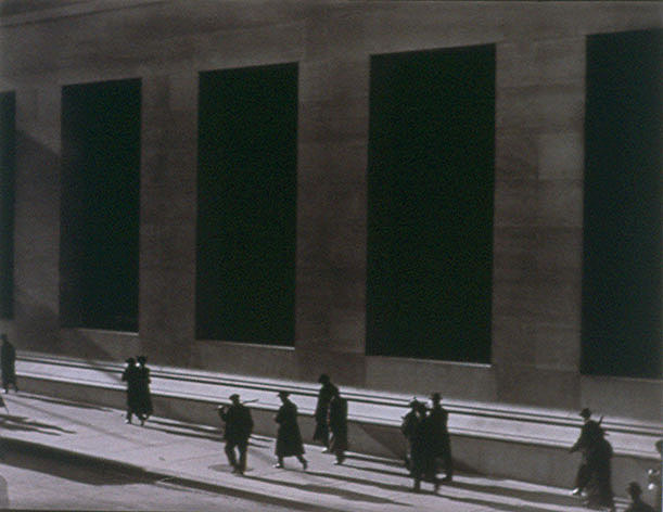 A black and white photograph of an early morning or late night street view of men and women walking next to a tall wall with very large long windows. Their shadows are cast long and thin.