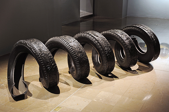 A stretched black tire in a spiral form, that is much longer than a normal tire—maybe five times the normal length. It has elaborate designs on the tread, sits on the floor, stretched out into five different circles and is illuminated by light.
