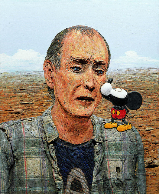A wooden carving that is painted of a middle aged man with grey hair, blue eyes that look shocked is wearing a flannel shirt and is being kissed by Mickey Mouse.