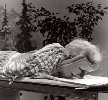 A black and white photograph of a patient wearing a padded hospital mask to cover injuries while lying face down on a trolley. They appear to be outdoors, surrounded by trees and plants and foggy mountains.