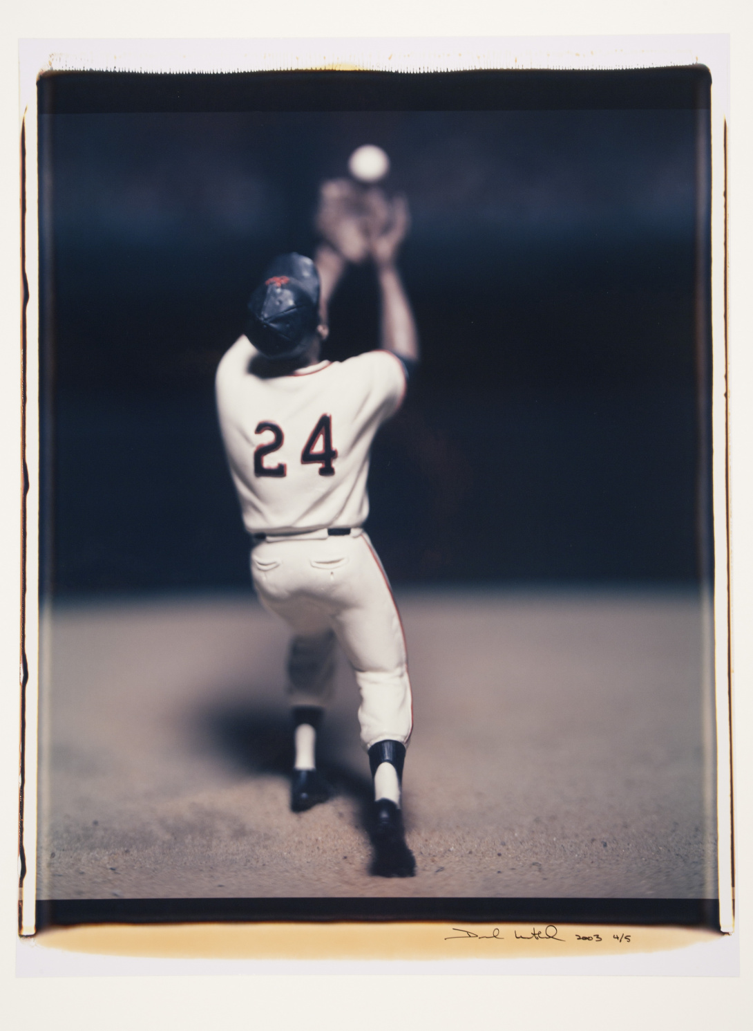 A photograph that shows the back of Willie Mays—he is wearing his jersey with #24 on it—in motion catching a baseball. The image is against a dark background and his arms, glove, and baseball are out of focus.