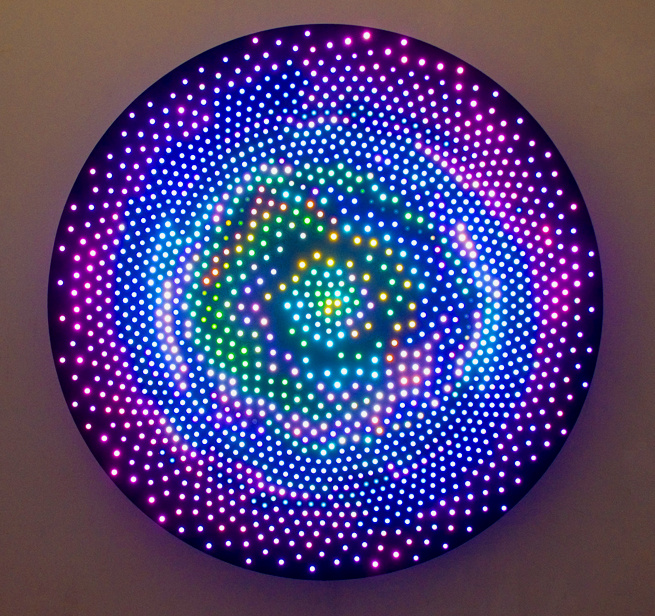 A round light sculpture is mounted onto a wall. The surface of the sculpture glows with hundreds of dots lit in varying hues of purple and blue to green and yellow at the center. The glowing lights are arranged in a circular shape which morphs into a flower towards the middle.