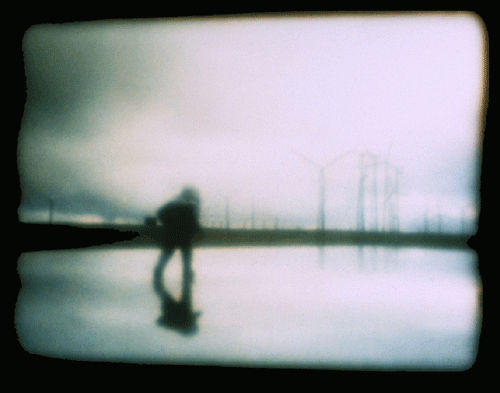 A blurry photo with windmills and a sun rising in the background. A nondescript person walks away from the viewer. He is wearing a heavy winter jacket and a hat to keep him warm from the cold weather. His blurry shadow reflects against the ground.