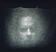 A photograph of a man's blurred face projected on a screen. It is fuzzy and his face is indistinct, perhaps in pain. The screen hangs from circular hooks against a black wall.