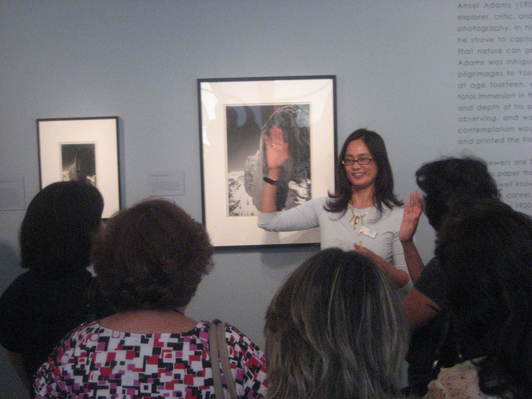 A docent-led tour in front of a group of older guests—the docent is presenting about a black and white photograph. One person in the crowd is raising their hand. 