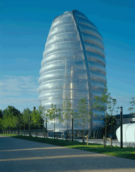 A photograph of a building with a spaceship shaped structure. It is wrapped in an outer layer of perforated metal that unites the building from afar. There are trees and grass surrounding the building, making it appear idyllic.