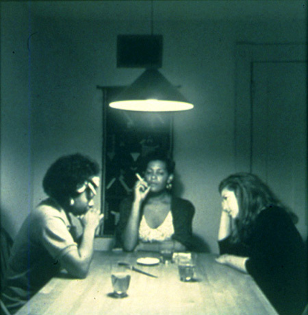 A black and white photograph of three women sit around a square table with a light hanging above them. They appear to be having a deep discussion and their style looks like 1970s or early 80s.