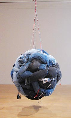 Installation of a large spherical object suspended by thin ropes only a few inches from the gallery floor. The sphere is constructed out of denim jeans that have been stuffed and looped together into a large ball. The denim jeans are black and various shades of blue. 