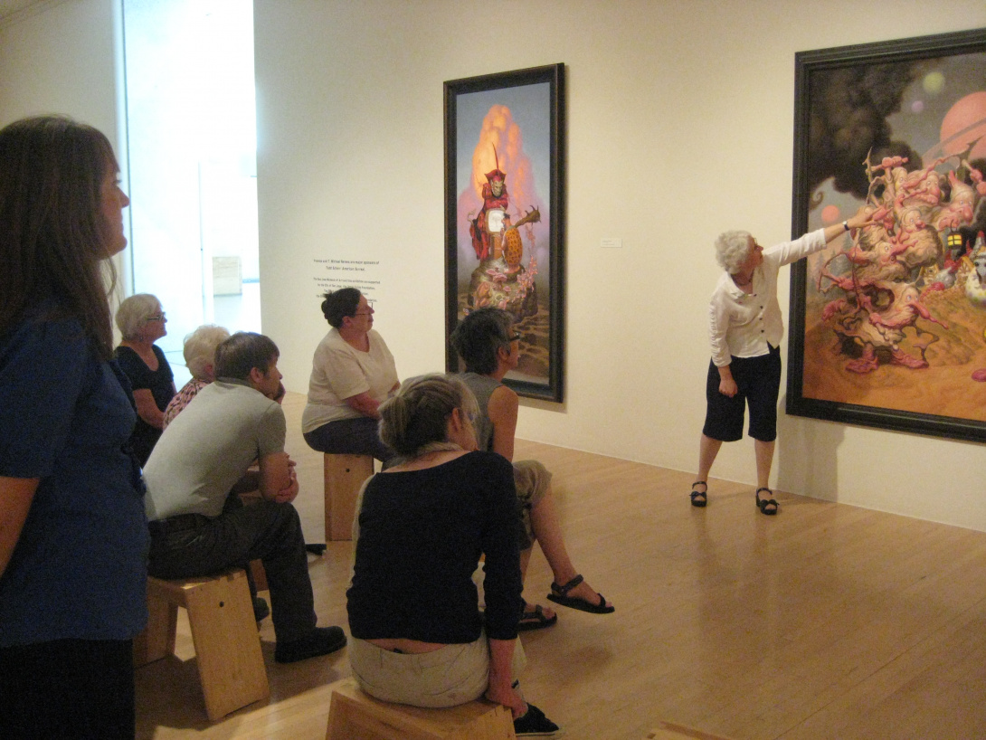 A docent leads a tour, she is standing and pointing towards a piece hanging on the wall. There is a group of people both standing and sitting on wooden chairs listening to the docent speak.