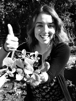 A woman in her late teens to early 20s photographed in black and white, showing a plant, still in punnet, to the viewer. She is smiling with her teeth and dimples showing and giving a 'thumbs up' gesture towards the viewer.