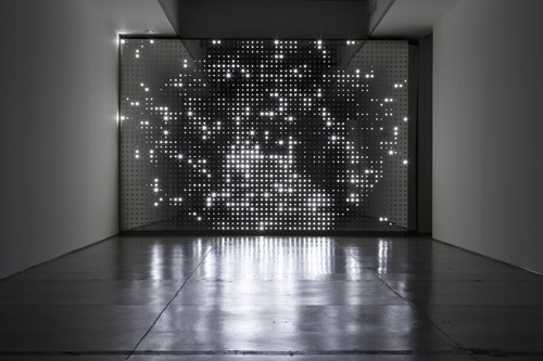 Installation of a glowing, metal screen that stretches the entire length of the room. The screen is lit from within or from behind and resembles an abstract map like a satellite image of a city from space. The space is dark and the lights reflect against the dark cement floor.
