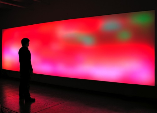 A visitor stands immersed in front a large, glowing screen that is is filled with a blurry sea of red, pink, green, and white. The image itself is indecipherable and resembles an abstract painting with a spray paint quality.