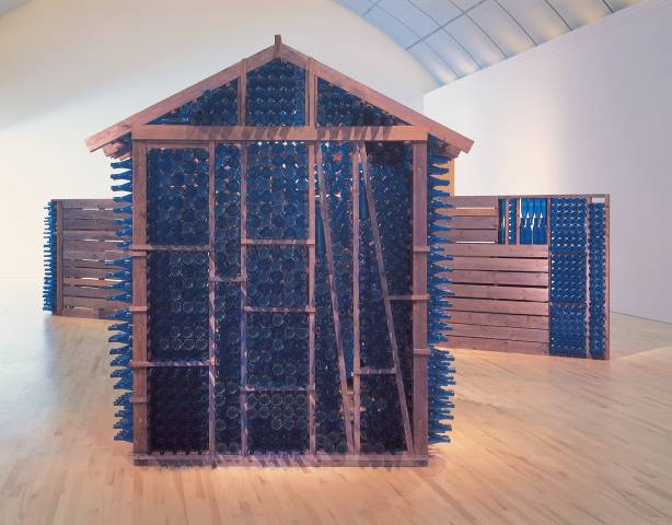 View of a gallery with 3 housing structures made of dark wood and cobalt glass bottles. The main structure is a one room building, large enough to stand in and a slanted roof. Fences extend from the sides of the building.