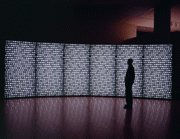 Six large screen panels side by side showing black and white bricks - almost mosaic-like with straight patterns on the top and more square-ish patterns below. There is also a man, lit from the screens, viewing the artwork.