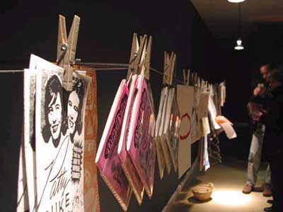 A dark room, with various zines hanging from a wire on clothespins. They are lit from above. There is a basket in front of them. In the distance, two people look at the zines.