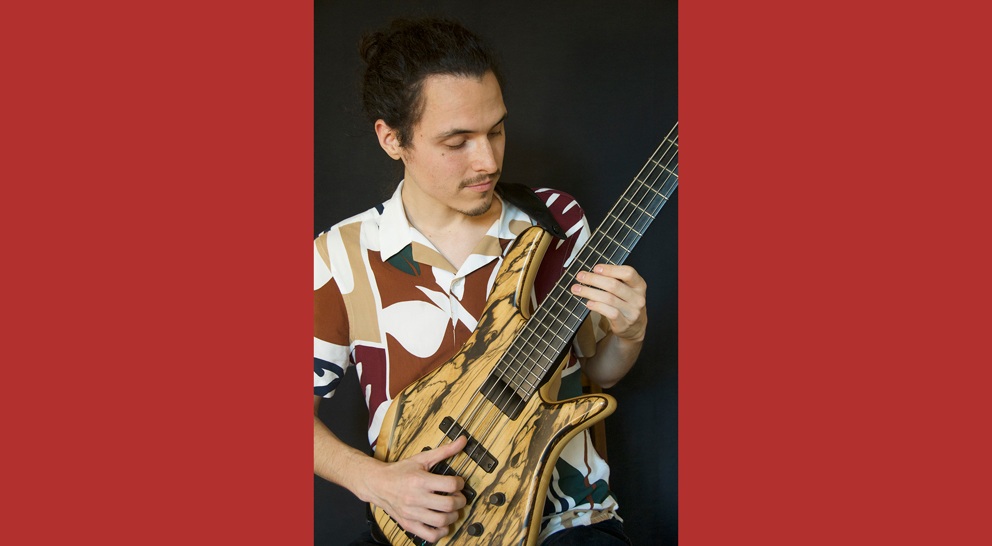 A man with a bun playing a six-string bass guitar with a natural wood finish, wearing a patterned shirt.