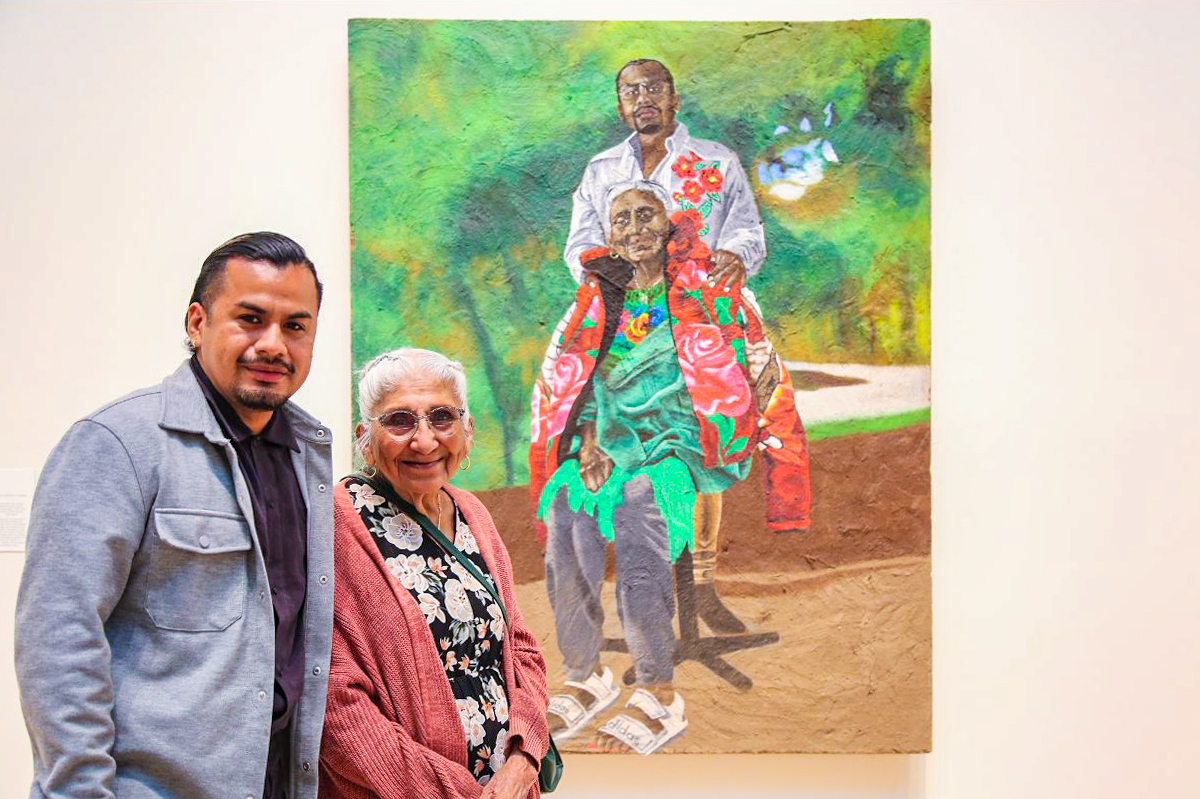 Yosimar Reyes and his grandmother, Mardonia Galena, in front of their painted portrait, which features Yosi standing behind his grandmother in an outdoor landscape.